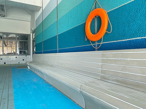 Bild von grauen Wärmebänken vor blauer Wand mit Orangenem Rettungsring in der Schwimmhalle Anton-Saefkow-Platz