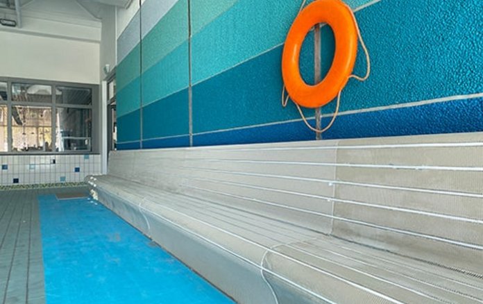 Bild von grauen Wärmebänken vor blauer Wand mit Orangenem Rettungsring in der Schwimmhalle Anton-Saefkow-Platz