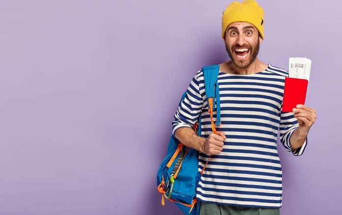 Mann mit gelber Mütze und Rucksack hält zwei Tickets hoch